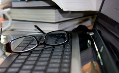 Grafika przedstawia laptopa , gdzie na klawiaturze położone są okulary. W tle widać stos książek